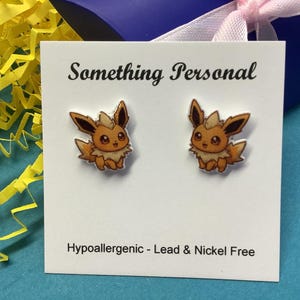 May include: A pair of stud earrings featuring a cartoon Eevee design. The earrings are on a white card with the text "Something Personal" and "Hypoallergenic - Lead & Nickel Free". The background includes yellow paper shreds and a blue surface.