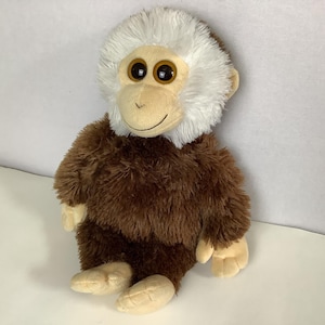 May include: A plush monkey toy with a brown body and a white head. The monkey has large, round eyes and tan hands and feet. The toy is sitting on a white surface.