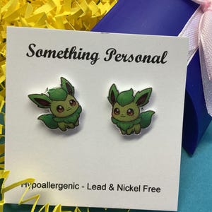 May include: A pair of green and brown cartoon character earrings on a white card with the text "Something Personal". The earrings are hypoallergenic and lead & nickel free. The earrings are on a white card, with yellow paper and a blue box in the background.