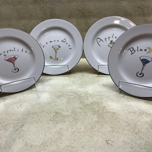 May include: Four white decorative plates with black rims, each featuring a different cocktail design and name: Cosmopolitan, Lemon Drop, Apple, and Blue Moon. The plates are displayed on metal stands.