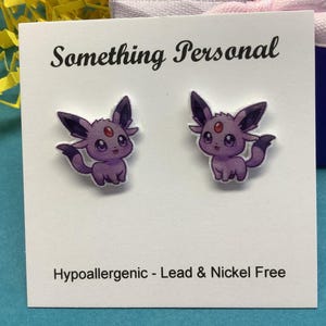 May include: A pair of purple cartoon character earrings. The earrings are on a white card with the text "Something Personal" and "Hypoallergenic - Lead & Nickel Free". The earrings feature a cute, smiling creature.