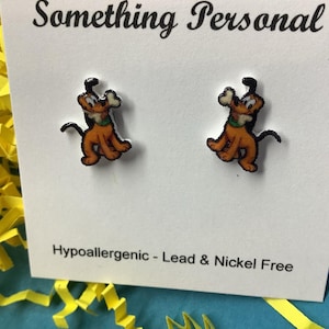 May include: A pair of cartoon dog stud earrings. The earrings feature a brown and orange dog with a bone in its mouth. The earrings are on a white card with the text "Something Personal" and "Hypoallergenic - Lead & Nickel Free".