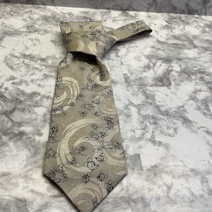 May include: A beige necktie featuring a repeating pattern of Mickey Mouse figures in black and white. The tie is knotted, showcasing the design's details. The background has a marble-like pattern.