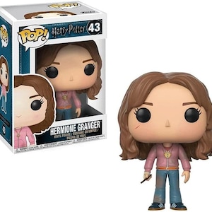 May include: A Funko Pop! vinyl figure of Hermione Granger from the Harry Potter series. The figure has a large head, black eyes, and brown hair. She wears a pink jacket, blue jeans, and holds a wand. The box is also visible.