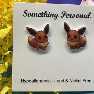 May include: A pair of Eevee stud earrings on a white card with the text "Something Personal". The earrings feature the cartoon character in brown, tan, and white. The card also states "Hypoallergenic - Lead & Nickel Free".