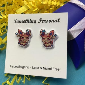 May include: A pair of colorful stud earrings featuring a cartoon character design. The earrings are on a white card with the text "Something Personal" and "Hypoallergenic - Lead & Nickel Free". A blue gift box with a pink ribbon is in the background.