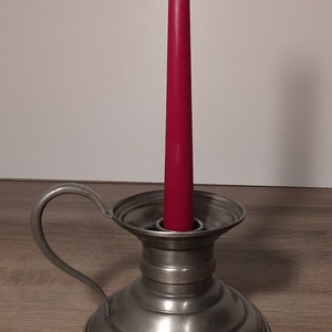 Può includere: Un candeliere in peltro con manico contiene una candela rossa alta e affusolata. Il candeliere ha una base rotonda e una parte superiore svasata. Il manico si curva verso l'alto dal lato. La candela è alta circa 20 cm.