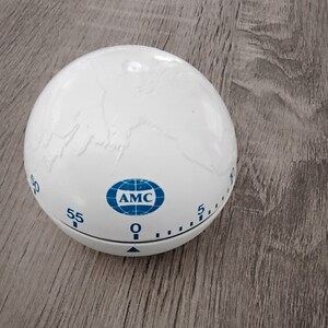 Peut inclure: Minuteur de cuisine blanc avec un design de globe et le texte "AMC" en bleu. Le minuteur a un cadran noir avec des chiffres de 5 à 60.
