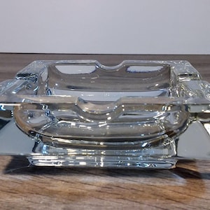 Könnte beinhalten: Ein klarer, quadratischer Aschenbecher aus Glas mit einem dicken, facettierten Design. Der Aschenbecher hat zwei Rillen zum Ablegen von Zigaretten und eine zentrale Vertiefung. Das Glas reflektiert das Licht und erzeugt einen Glanzeffekt.