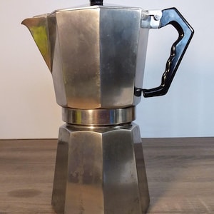 May include: A silver metal stovetop espresso maker with a black plastic handle and a black plastic lid.