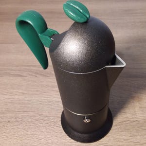 Op de afbeelding: Een zwarte en groene espressomachine voor op het fornuis met een koffieboonvormige knop op de deksel. De machine heeft een groene handgreep.
