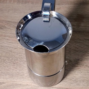 Op de afbeelding: Een roestvrijstalen espressomaker, ook wel een Moka pot genoemd, met een scharnierend deksel en een handvat. De pot heeft een cilindrisch lichaam en een glanzend, reflecterend oppervlak.