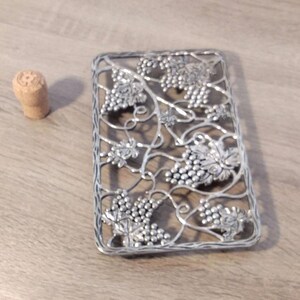May include: Silver-toned metal hot pad with a grapevine design. The pad is square shaped and has a decorative, intricate design.