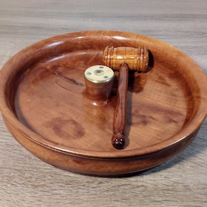 Può includere: Un martelletto di legno, un piccolo cilindro con la parte superiore in ottone e una ciotola di legno. Il martelletto ha una testa arrotondata e un lungo manico. La ciotola è rotonda con un bordo largo. Il cilindro è piccolo e cilindrico. Tutti gli oggetti sono in legno.