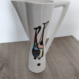 May include: White ceramic pitcher with a stylized bird design in black, red, yellow, blue, and green. The pitcher has a flared spout and a handle.
