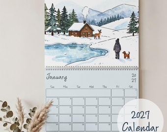 Puede incluir: Un calendario de pared para 2027 con una ilustración en acuarela de un paisaje invernal nevado con una cabaña de madera, pinos, un estanque helado, un ciervo y una persona paseando a un perro. La página de enero muestra una cuadrícula mensual.