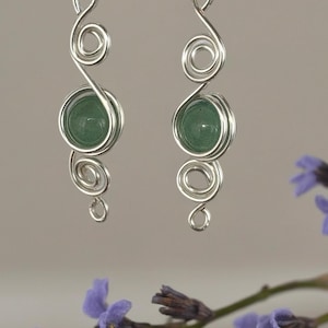 Puede incluir: Un par de pendientes colgantes plateados. Cada pendiente presenta una piedra preciosa verde engarzada en un diseño de espiral de alambre. Los pendientes tienen una parte superior en bucle para colgar y un remolino decorativo debajo.