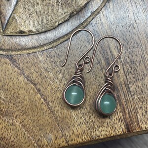 May include: A pair of copper wire wrapped earrings with green stone beads. The earrings are hanging from a wooden surface.