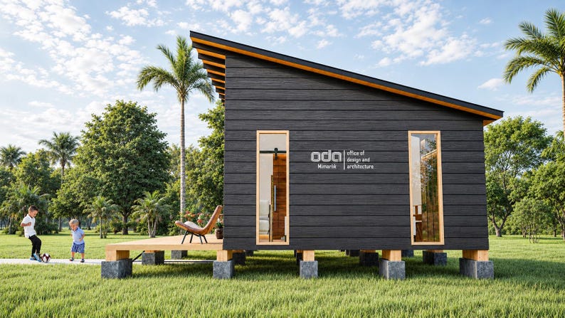 May include: A small, dark-sided cabin with a slanted roof, elevated on concrete block piers. The exterior features horizontal wood siding, two tall rectangular windows, and a wooden deck. The text "oda office of design and architecture" is visible.
