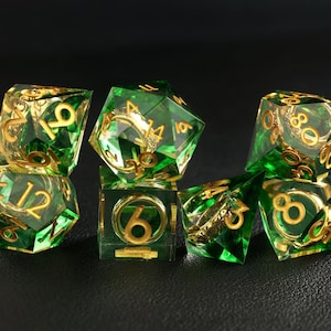 May include: Set of seven transparent green polyhedral dice with gold-colored numbers and accents. The dice are arranged on a dark surface, showcasing their multifaceted design. Ideal for tabletop gaming.