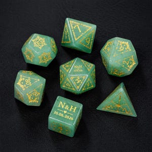 May include: A set of seven green stone polyhedral dice with gold-coloured engraved numbers and decorative filigree patterns. Two dice feature the custom text N&H and the date 10.06.2026. The set includes various geometric shapes for gaming.