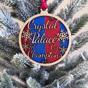 May include: A wooden Christmas bauble with the text "Crystal Palace FA Cup Champions 2025." The bauble features a red and blue glitter design, snowflake details, and a red ribbon for hanging on a Christmas tree.