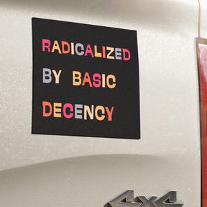 May include: A black rectangular magnet with the words "RADICALIZED BY BASIC DECENCY" in colorful block letters is affixed to the back of a white vehicle. The vehicle has a 4x4 emblem.