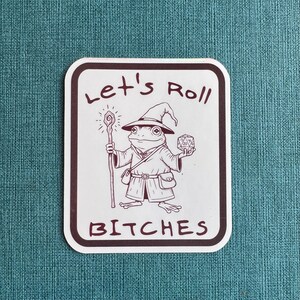 Pode incluir: Um autocolante branco com uma borda preta com um sapo de desenho animado vestido de mago, segurando uma bengala e um dado. O texto "Let's Roll BITCHES" está escrito no autocolante.