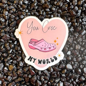 Puede incluir: Pegatina rosa en forma de corazón con el texto "You Croc My World" en una fuente caprichosa. Un zapato Croc rosa y blanco aparece en el centro del corazón.
