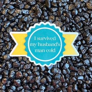 May include: A sticker with the text "I survived my husband's man cold" in white lettering. The sticker has a teal blue circle with a white scalloped edge and yellow banner accents. The background is a bed of dark, shiny stones.