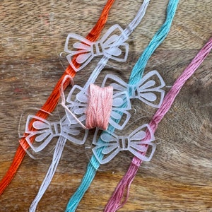 Könnte beinhalten: Vier transparente, plastik-geformte Schleifen-Faden-Organizer mit Spulen von rosa, orange, blau und rosa Garn.