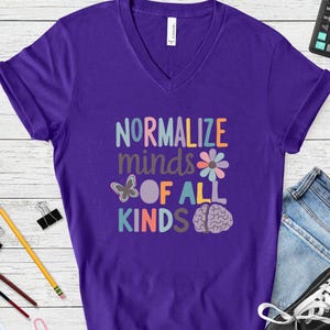 May include: A purple V-neck t-shirt featuring the text "NORMALIZE minds OF ALL KINDS" in multi-coloured lettering. The design includes a small butterfly, a stylised flower, and a brain illustration in soft pastel tones printed on the front centre.