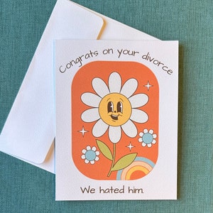 Könnte beinhalten: Eine Grußkarte mit einer Comic-Gänseblümchen mit einem fröhlichen Gesicht. Die Karte hat einen orangenen Hintergrund mit einem Regenbogen und dem Text "Herzlichen Glückwunsch zur Scheidung. Wir haben ihn gehasst."