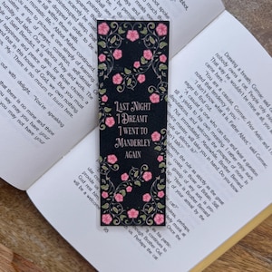 May include: Black bookmark with pink floral accents and the text "LAST NIGHT I DREAMT I WENT TO MANDERLEY AGAIN." The bookmark is rectangular and is placed in an open book.