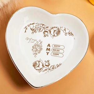 May include: Heart-shaped white ceramic dish with a gold rim. The dish features a floral design and the name "AMY" with the words "ACCEPTED", "MADE NEW", and "YEARNING". The phrase "God says, I am" is also printed on the dish.