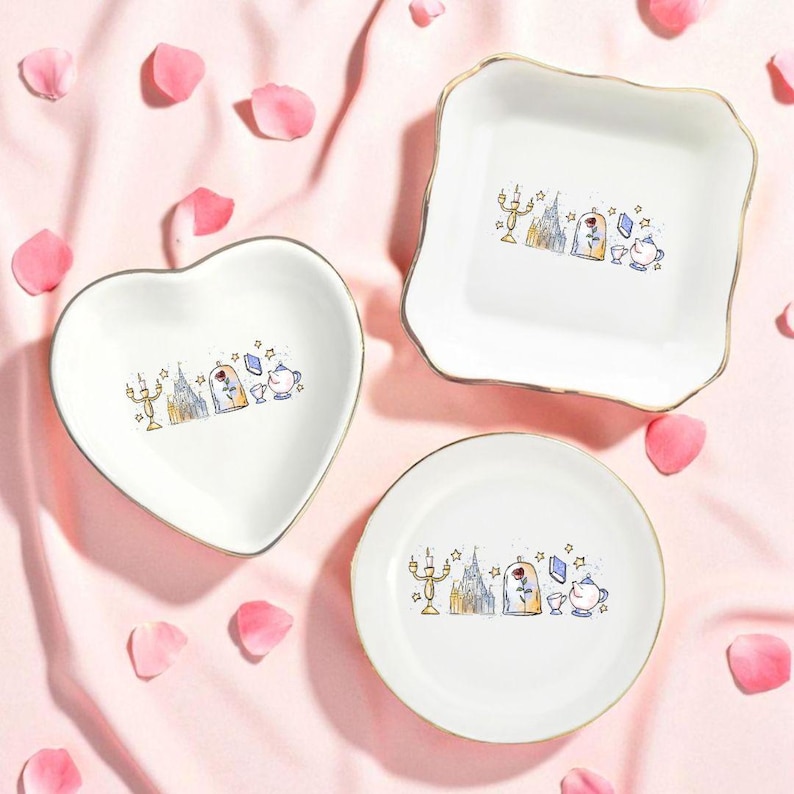 May include: Three white ceramic dishes in heart, square, and round shapes, each with a gold rim. The dishes feature a colorful illustration of a castle, rose, and teapot. Pink rose petals are scattered around the dishes on a pink fabric background.