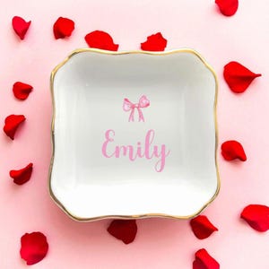 K&ouml;nnte beinhalten: Eine wei&szlig;e quadratische Schmuckschale mit goldenem Rand, mit einer rosa Schleife und dem Namen "Emily" in rosa Schrift. Rote Rosenbl&auml;tter sind um die Schale auf einem rosa Hintergrund verstreut. Ein dekorativer und personalisierter Artikel.
