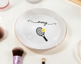 May include: White ceramic trinket dish with a gold rim. The dish features a black tennis racket graphic with a yellow tennis ball and the name "amy" in cursive. Makeup brushes are visible in the background.