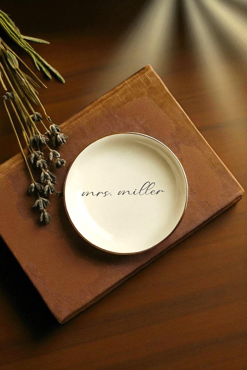 May include: A small, off-white ceramic dish with a gold rim, inscribed with the name "mrs. miller" in black script. The dish rests on a brown book, with dried lavender sprigs nearby. A simple, elegant design.