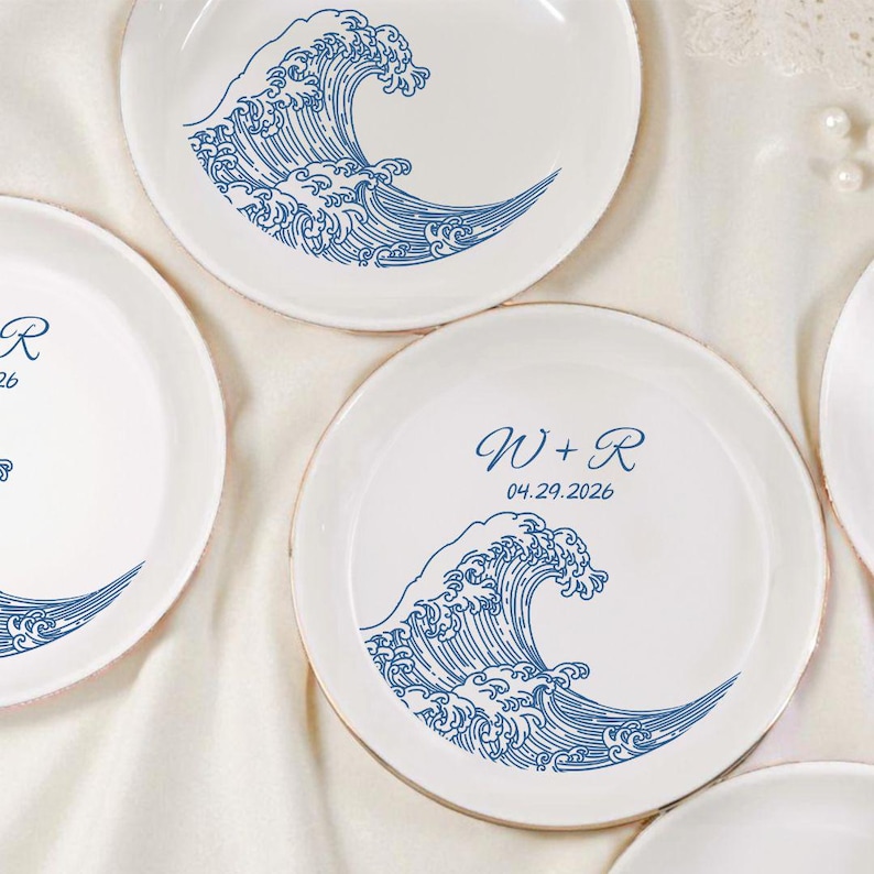 May include: White ceramic plates with a blue wave design. Some plates feature initials and a date. The wave design is a stylized illustration. The plates have a slightly textured rim.