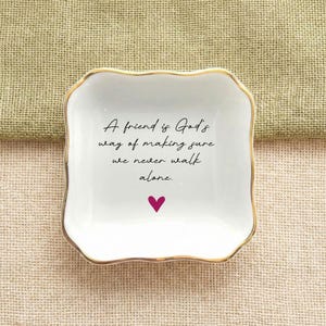 May include: A square white ceramic trinket dish with scalloped edges and a gold-toned rim. The center features black cursive text that reads, A friend is God's way of making sure we never walk alone, followed by a small solid pink heart icon.