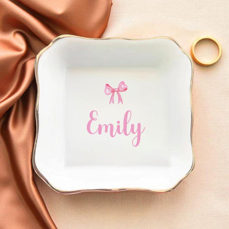 K&ouml;nnte beinhalten: Eine wei&szlig;e Keramik-Schmuckschale mit goldenem Rand und gewellten Kanten. Die Schale zeigt eine rosa Schleifen-Grafik und den Namen "Emily" in rosa Schrift. Ein goldener Ring liegt im Hintergrund.