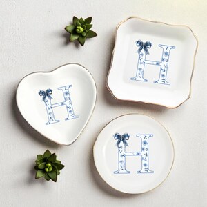 May include: A collection of three white ceramic trinket dishes in heart, square, and round shapes. Each dish features a blue floral letter "H" design with a bow. The square and round dishes have gold rims. Green succulents are placed around the dishes.