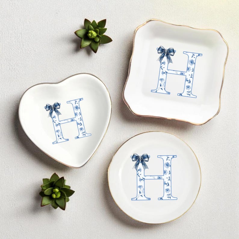 May include: A collection of three white ceramic trinket dishes in heart, square, and round shapes. Each dish features a blue floral letter "H" design with a bow. The square and round dishes have gold rims. Green succulents are placed around the dishes.
