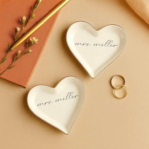 May include: Two heart-shaped white ceramic ring dishes with gold trim, each inscribed with "mrs. miller" in black script. A gold pen and a small notebook are visible in the background, along with two gold rings.