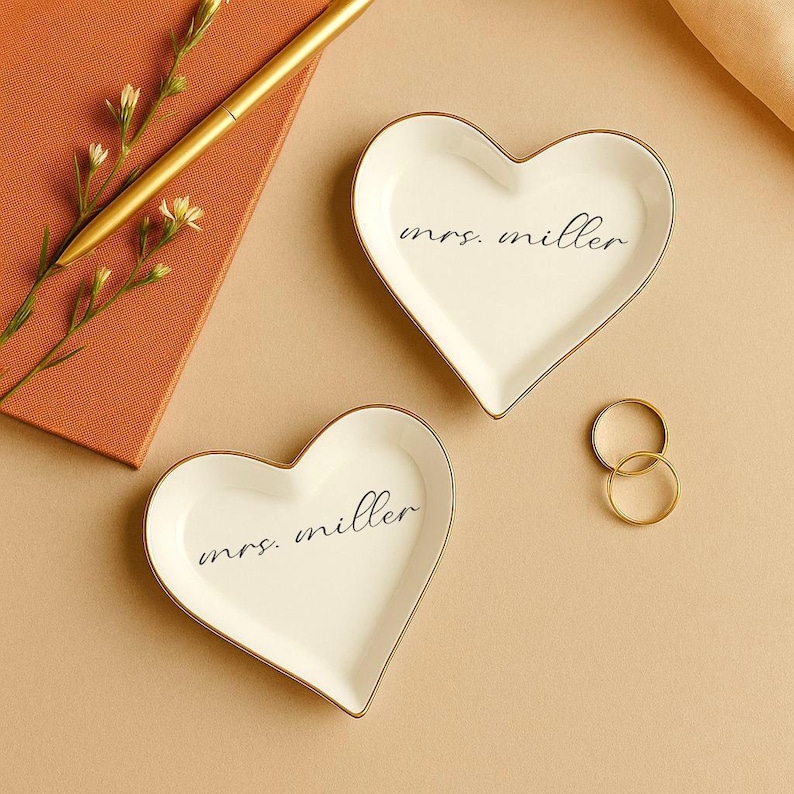 May include: Two heart-shaped white ceramic ring dishes with gold trim, each inscribed with "mrs. miller" in black script. A gold pen and a small notebook are visible in the background, along with two gold rings.