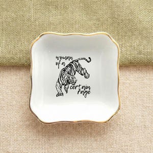 Peut inclure: Un vide-poche carré en céramique blanche avec un bord doré. Le plat présente une illustration noire d'un tigre et le texte "woman of a certain rage". Le plat a un bord ondulé.