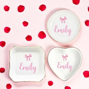 K&ouml;nnte beinhalten: Drei wei&szlig;e Keramik-Schmuckschalen mit goldenen R&auml;ndern, jeweils mit einer rosa Schleife und dem Namen "Emily". Eine ist rund, eine quadratisch und eine herzf&ouml;rmig. Rote Rosenbl&auml;tter sind auf einem rosa Hintergrund verstreut.