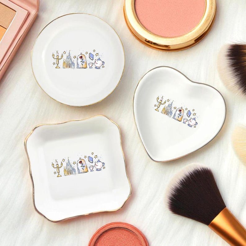 May include: White ceramic trinket dishes in round, heart, and square shapes, each with a gold rim. The dishes feature a colorful illustration of a castle, rose, and teacup. Makeup brushes and blush are in the background.