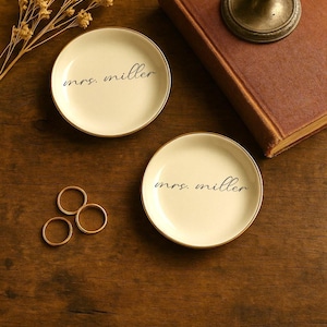 May include: Two small, off-white ceramic dishes with gold rims, each inscribed with "mrs. miller" in black script. Three gold rings and a vintage candlestick holder sit on a wooden surface, alongside a closed book and dried flowers.